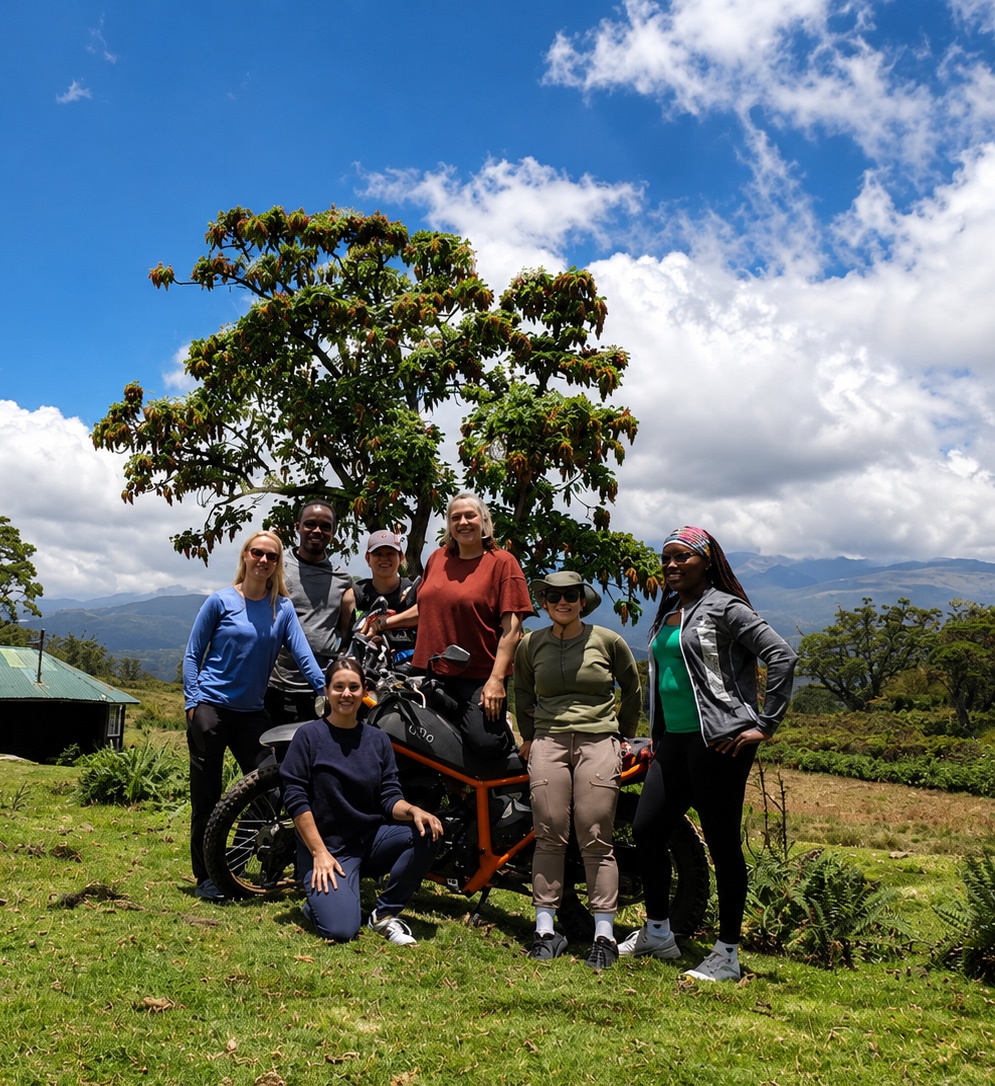 Group of six people posing outdoors in a lush green field with a large tree and a rustic building in the background, under a bright blue sky with scattered clouds.