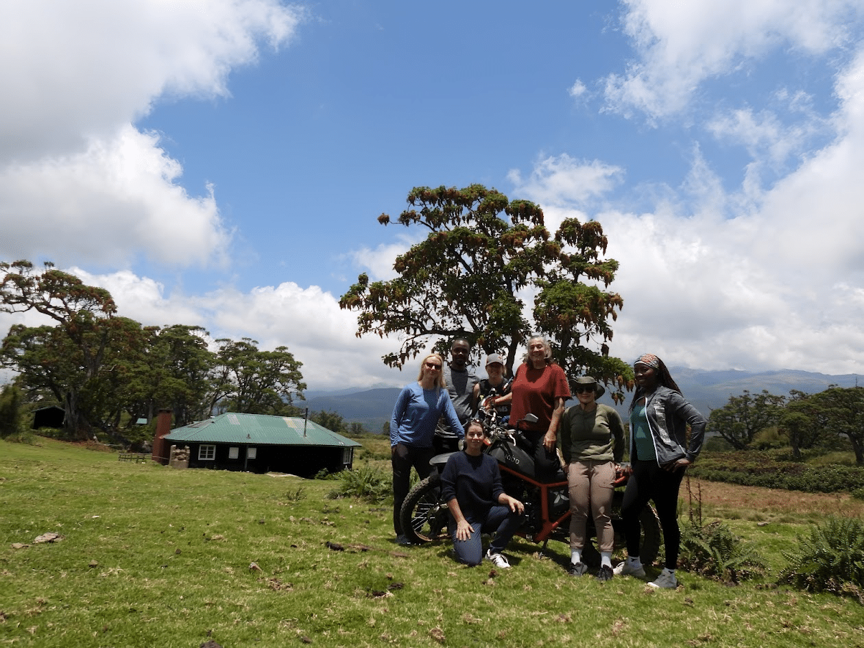 Group of six people posing outdoors in a lush green field with a large tree and a rustic building in the background, under a bright blue sky with scattered clouds.
