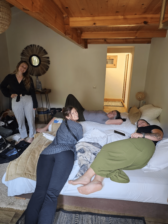 Group of friends relaxing on a bed in a cozy room, with one person standing nearby and others lounging comfortably. Natural light fills the space, highlighting a casual and friendly atmosphere.