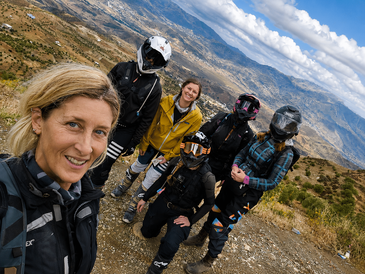 Group of five adventure enthusiasts posing for a selfie on a mountain trail, wearing motorcycle gear and helmets, with a scenic landscape in the background.