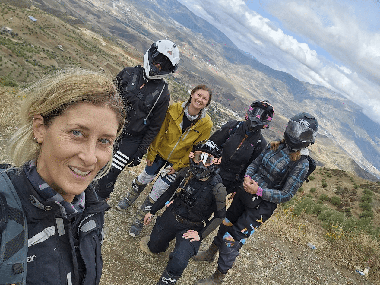 Group of five adventure enthusiasts posing for a selfie on a mountain trail, wearing motorcycle gear and helmets, with a scenic landscape in the background.