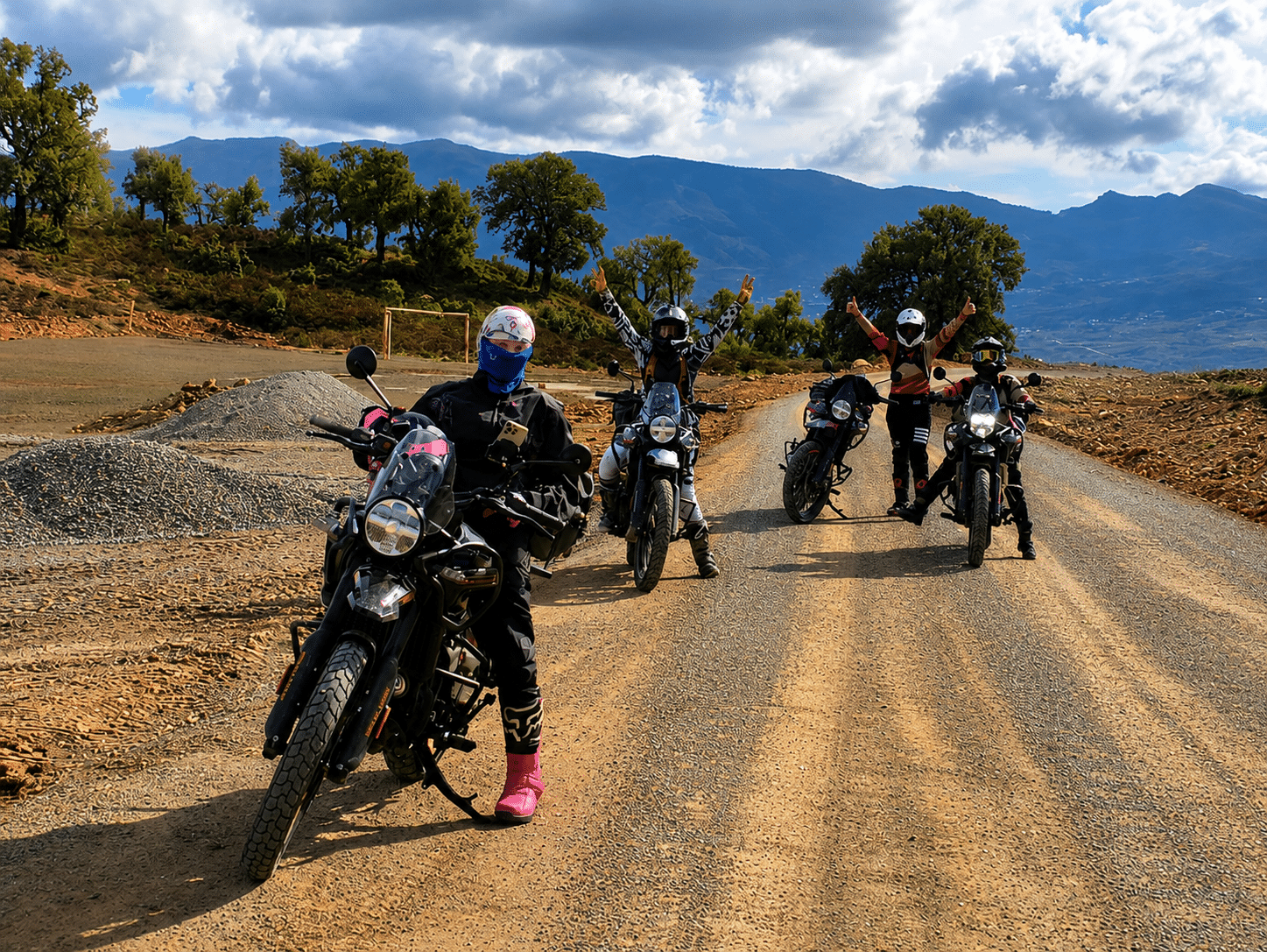Group of four motorcyclists posing on a dirt road surrounded by mountains, showcasing their adventure gear and motorcycles. The scene captures the spirit of off-road riding and camaraderie among enthusiasts.