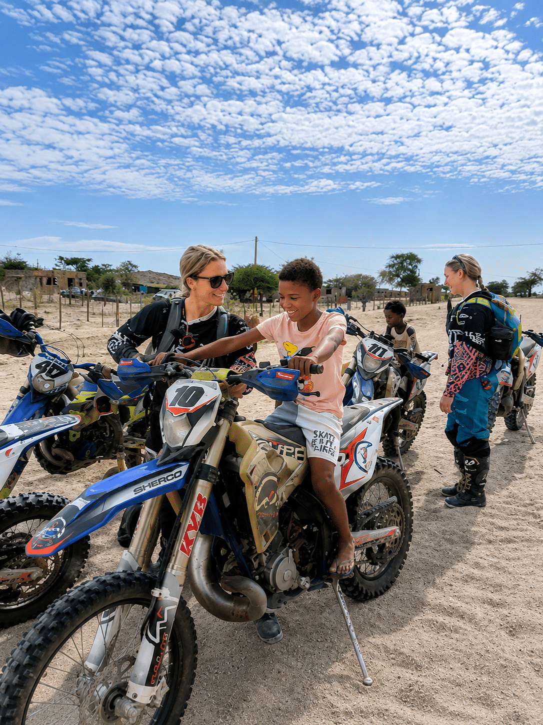 A group of individuals, including children and adults, engage in dirt biking activities in a sandy outdoor setting. One child is seated on a motorcycle while an adult assists them, showcasing a supportive environment for learning and adventure in off-road biking.