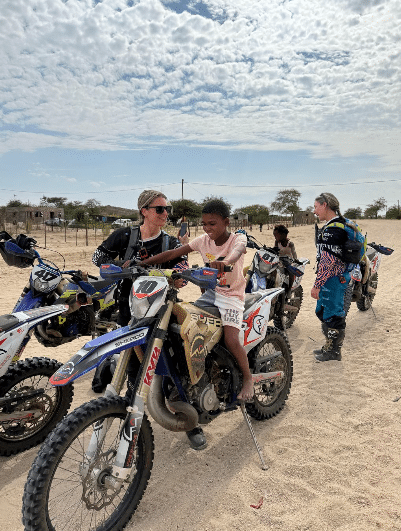 A group of individuals, including children and adults, engage in dirt biking activities in a sandy outdoor setting. One child is seated on a motorcycle while an adult assists them, showcasing a supportive environment for learning and adventure in off-road biking.