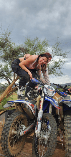 Two individuals posing on dirt bikes outdoors, showcasing a fun and adventurous atmosphere in a natural setting with trees and a cloudy sky.