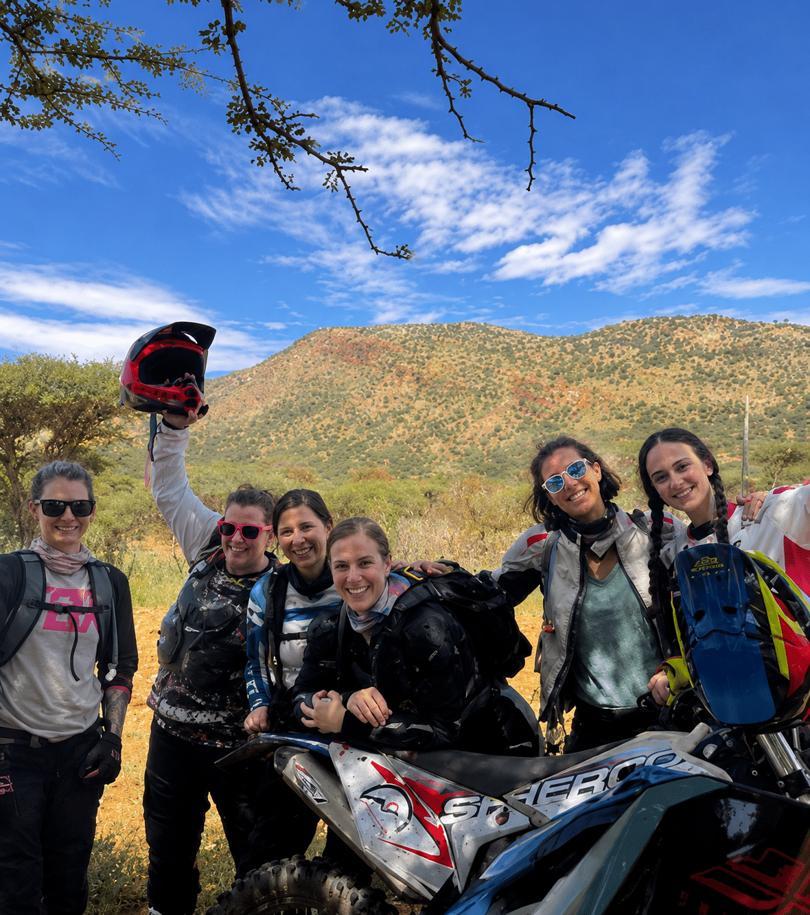 Group of six female motocross riders posing with their dirt bikes in a scenic outdoor setting, showcasing a mountainous landscape under a blue sky.