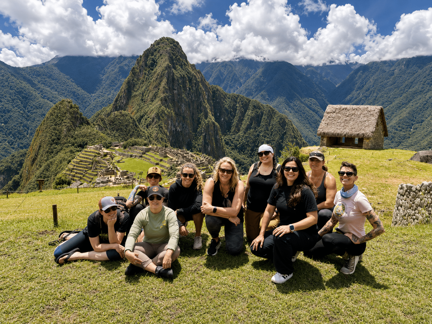 Group of travelers posing at Machu Picchu with mountains and ancient ruins in the background under a partly cloudy sky.