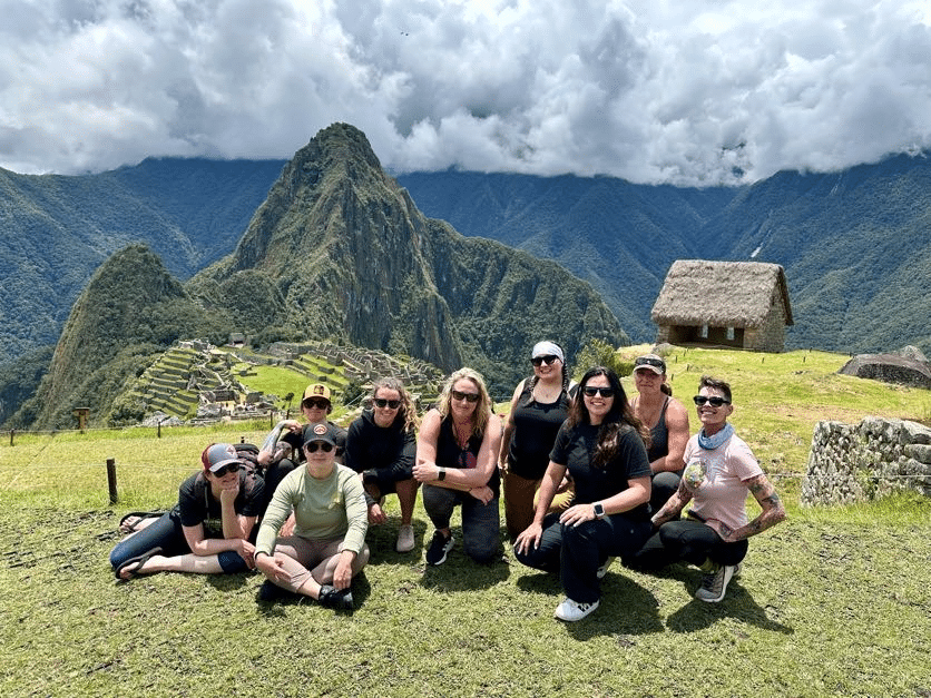 Group of women posing at Machu Picchu with mountain and ancient ruins in the background under a cloudy sky.