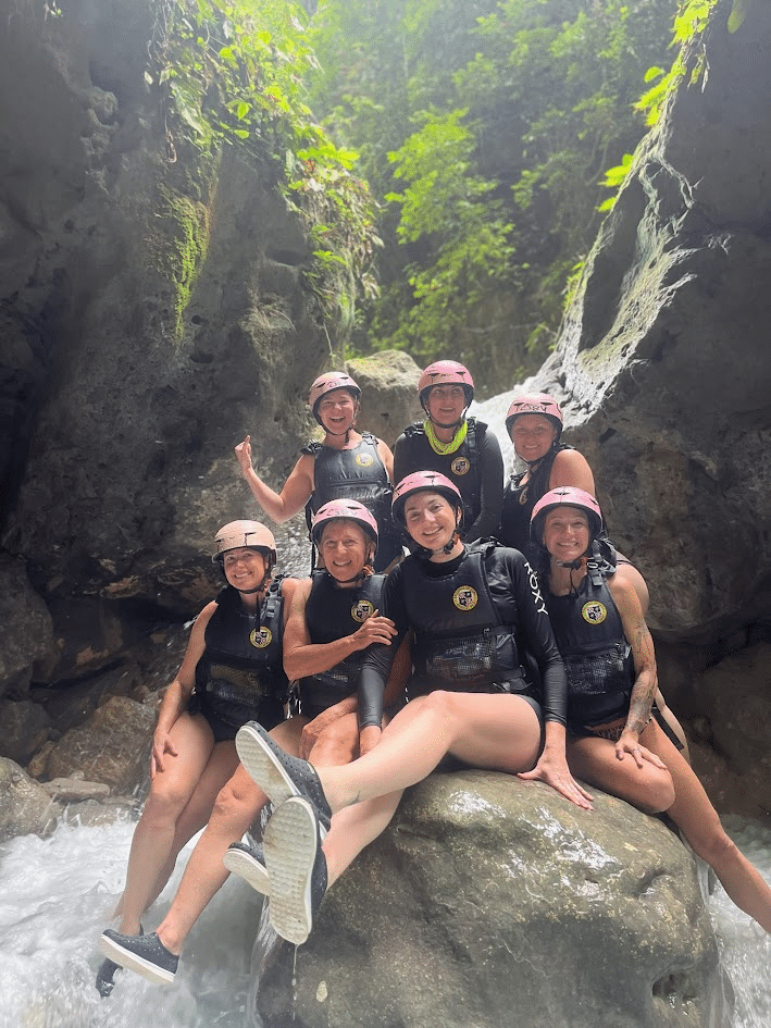 Group of seven women in wetsuits and helmets enjoying a river adventure, posing on rocks near a waterfall in a lush, green environment.