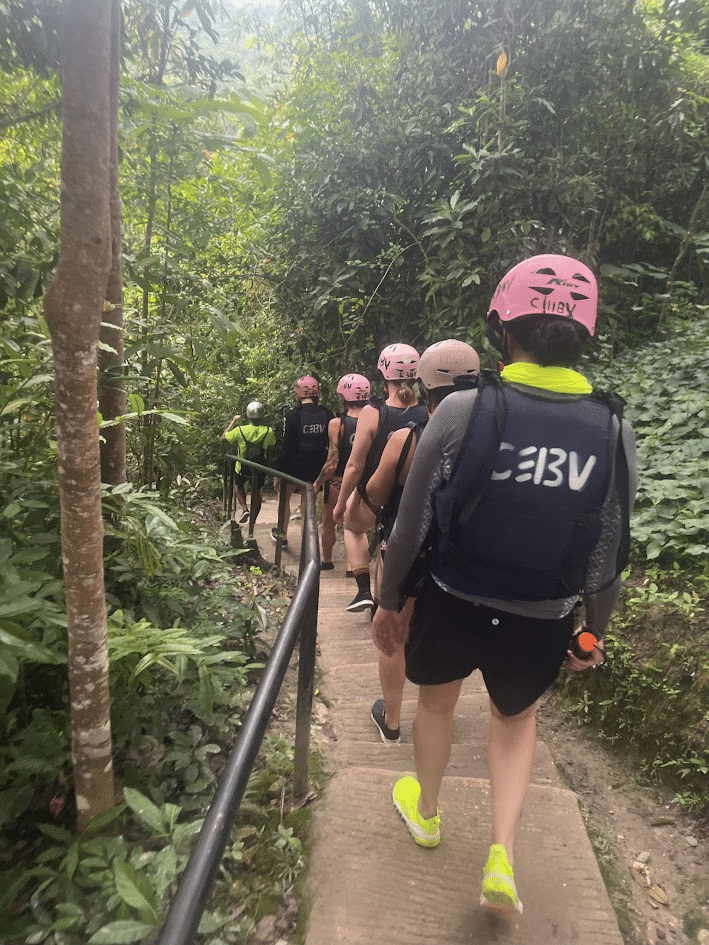 Group of adventure seekers hiking along a jungle trail, wearing helmets and life vests, preparing for an outdoor activity in a lush green environment.