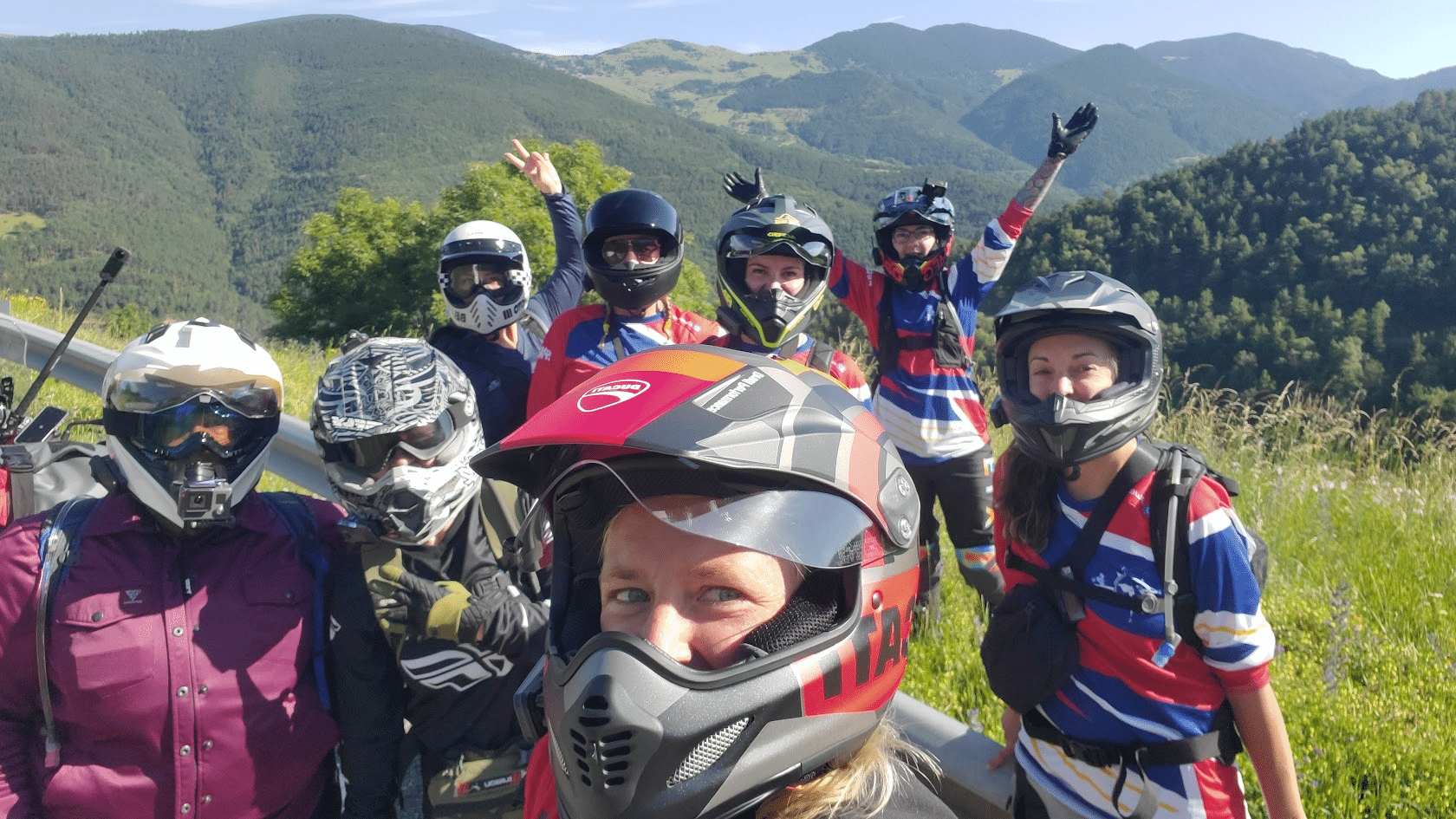 Group of women motorcyclists wearing helmets and gear taking a selfie in a mountainous outdoor setting with green hills and blue sky.