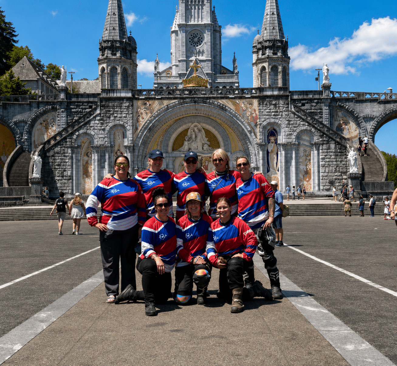 Group of people wearing matching red and blue sports jerseys posing in front of the Basilica of Our Lady of the Rosary in Lourdes, France.