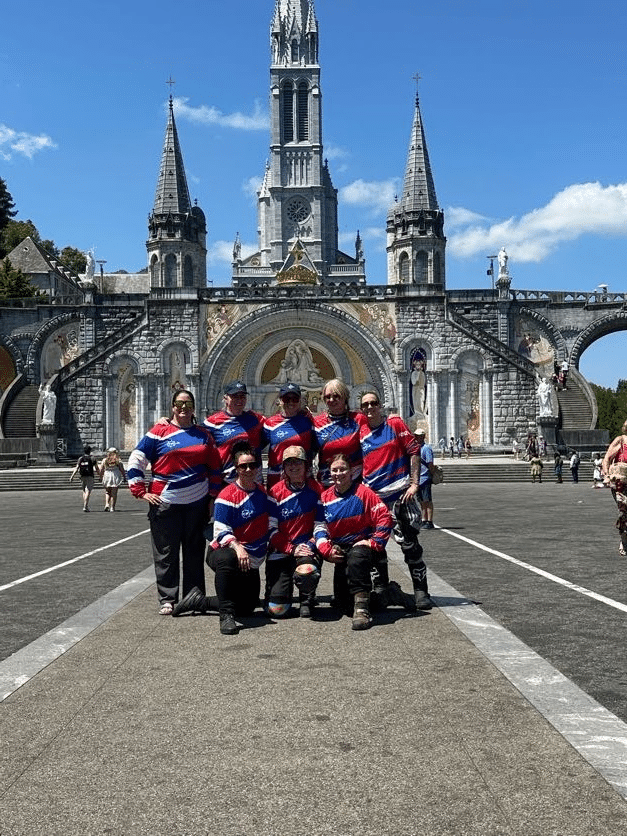 Group of women wearing matching red and blue sports jerseys posing in front of the Basilica of Our Lady of the Rosary in Lourdes, France on a sunny day.