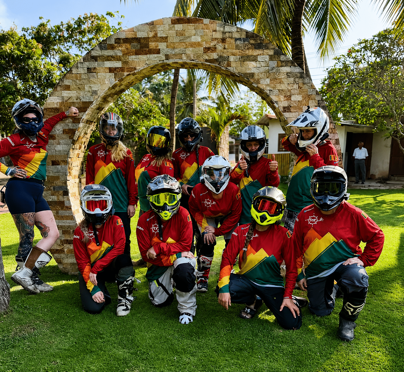 Group of motocross riders wearing helmets and matching red, green, and yellow jerseys posing outdoors in front of a stone arch.