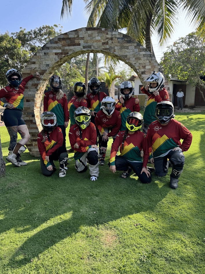 Group of motocross riders in colorful gear and helmets posing outdoors in front of a stone arch on a sunny day