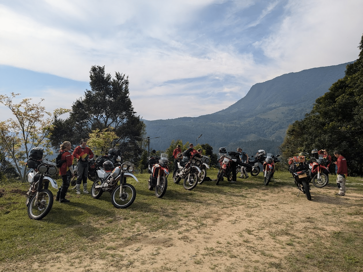 Group of motorcyclists with dual-sport bikes parked on a grassy hilltop with forested mountains and partly cloudy sky in the background.