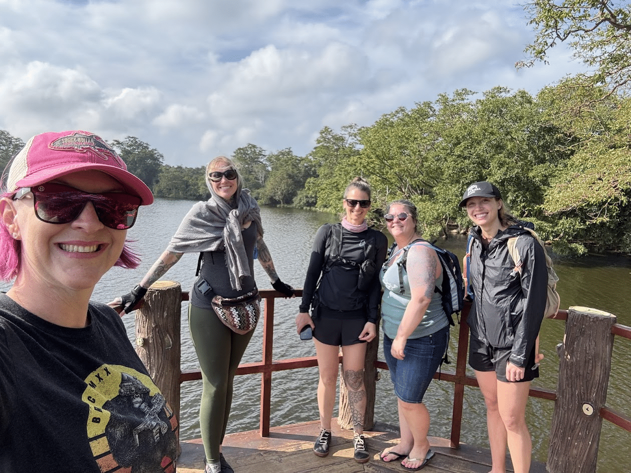 Group of five women wearing casual and outdoor clothing standing on a wooden platform by a river with trees and cloudy sky in the background.