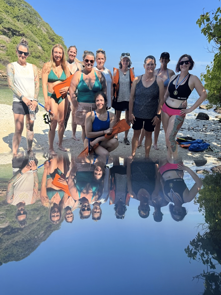 Group of women in swimsuits and casual wear standing on a beach with rocky shoreline and green hills, reflected in calm water under a clear blue sky.