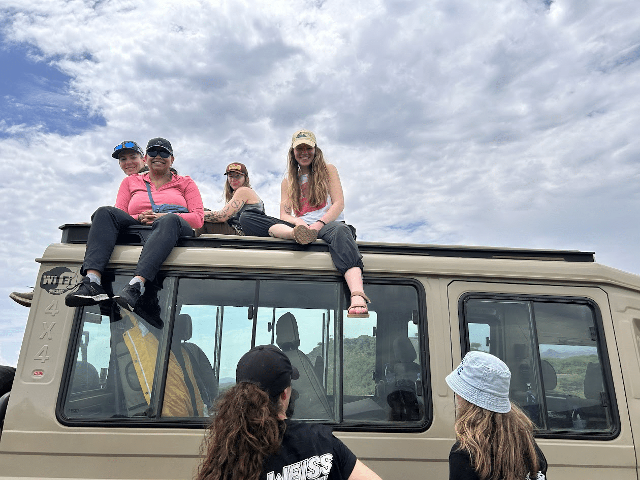 Group of friends enjoying an outdoor adventure, sitting on top of a 4x4 vehicle under a cloudy sky, showcasing a fun and relaxed atmosphere.