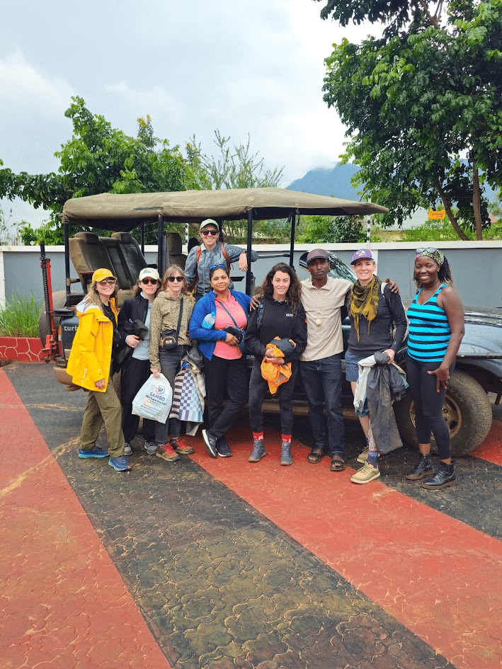 Group of travelers posing in front of a safari vehicle, surrounded by greenery and mountains, preparing for an adventure trip. The diverse group is dressed in outdoor gear, showcasing a spirit of exploration and camaraderie.