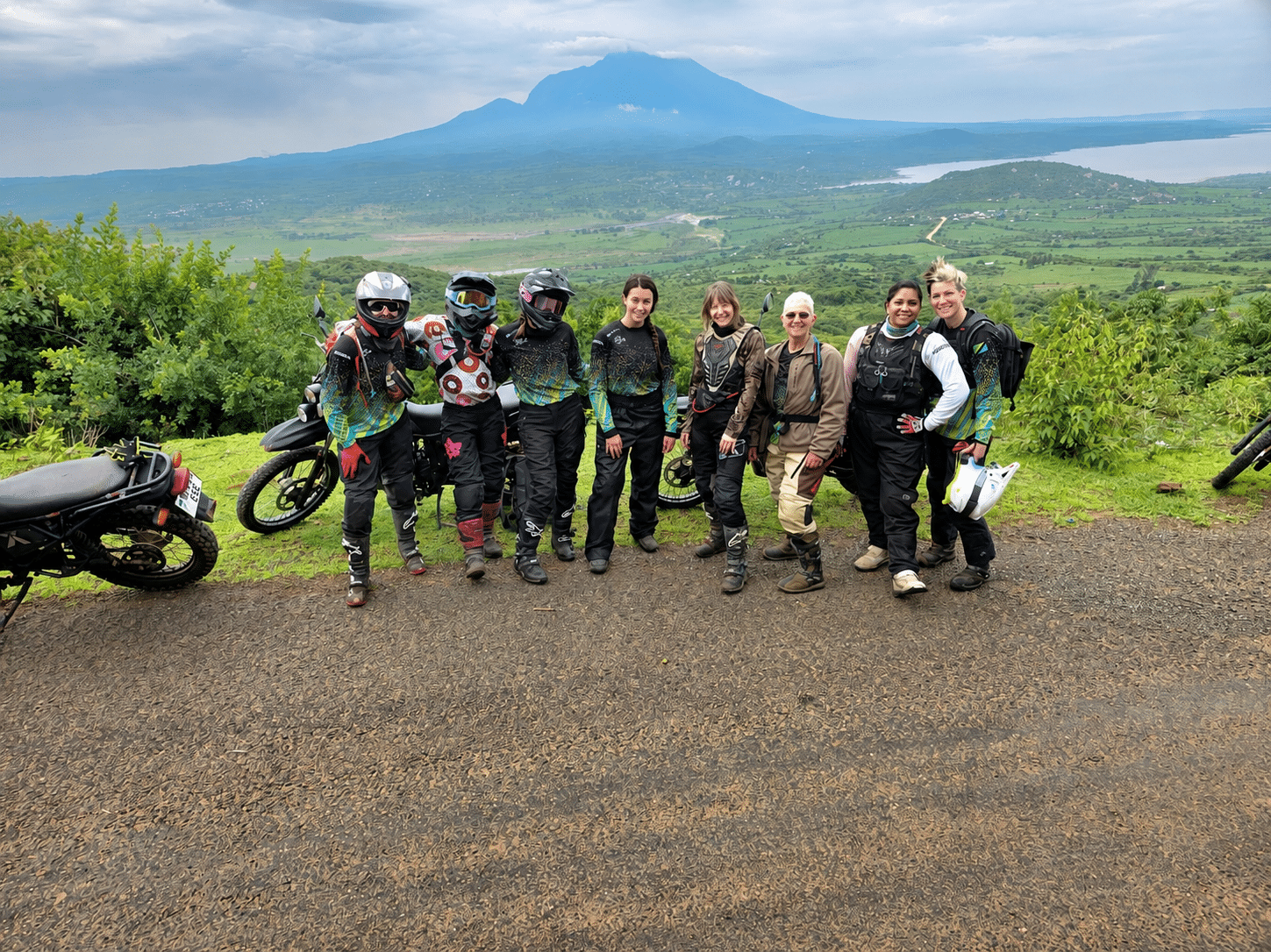 Eight women in motorcycle gear standing at a viewpoint with a large mountain in the background.