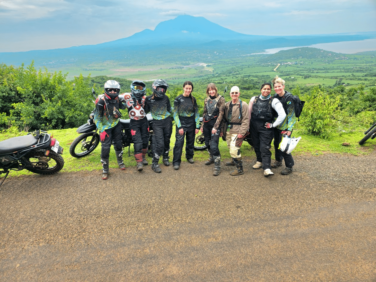 Eight women in motorcycle gear standing at a viewpoint with a large mountain in the background.