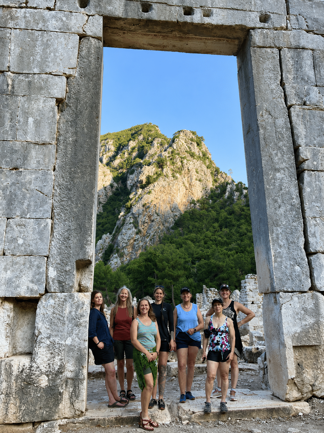 Group of seven women posing under ancient stone archway with mountainous landscape in the background during daytime.