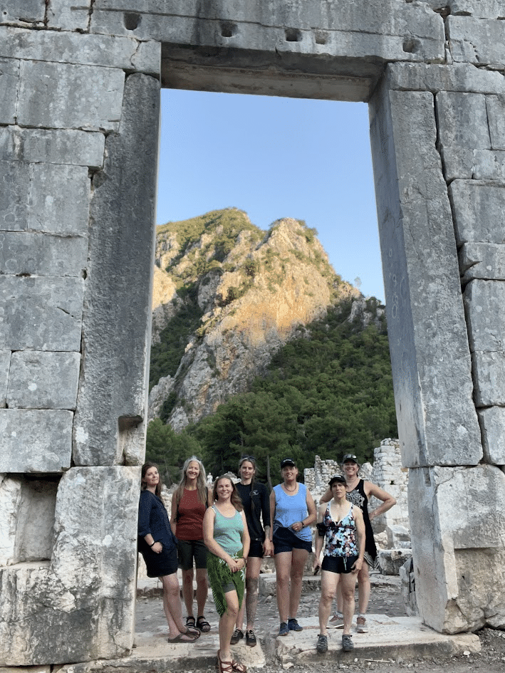 Group of women standing under large ancient stone archway with mountainous landscape in the background.