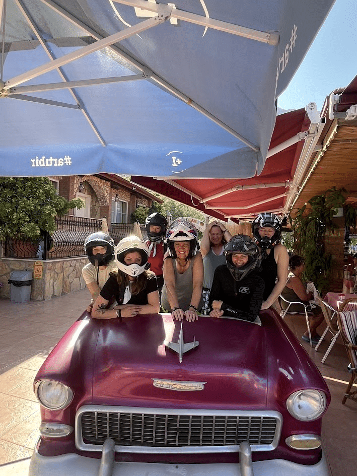 Group of women wearing motorcycle helmets posing behind a purple vintage car hood under outdoor umbrellas at a cafe.