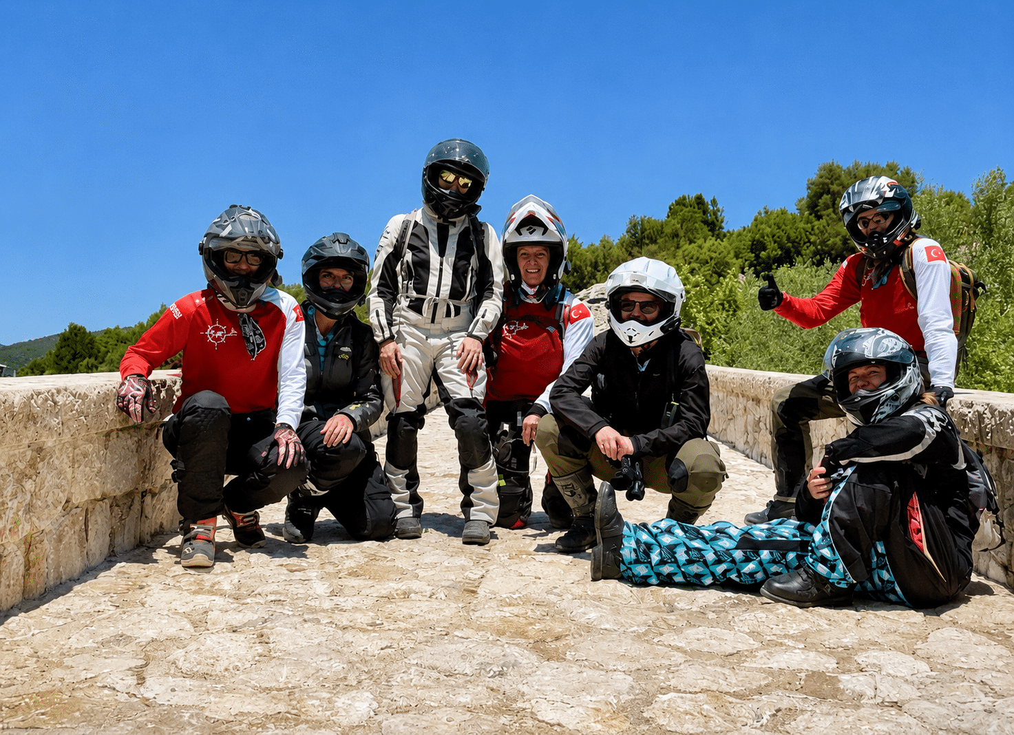 Group of seven women motorcyclists in full gear and helmets posing on a stone bridge under clear blue sky with green trees in the background.