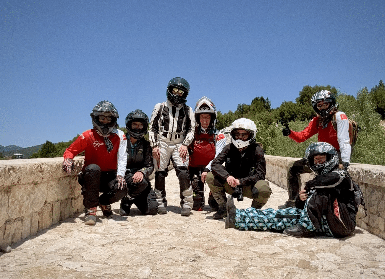 Group of seven motorcyclists wearing helmets and protective gear posing outdoors on a stone pathway with trees and clear blue sky in the background.