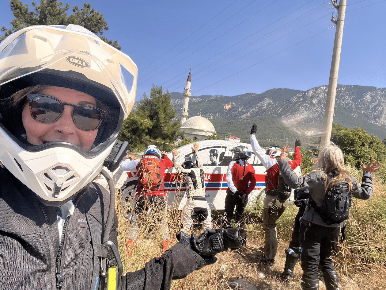 Rescue team members in protective gear gather near a vehicle in a mountainous area, with a mosque in the background. The image features a person in a helmet taking a selfie, highlighting their involvement in emergency response efforts.