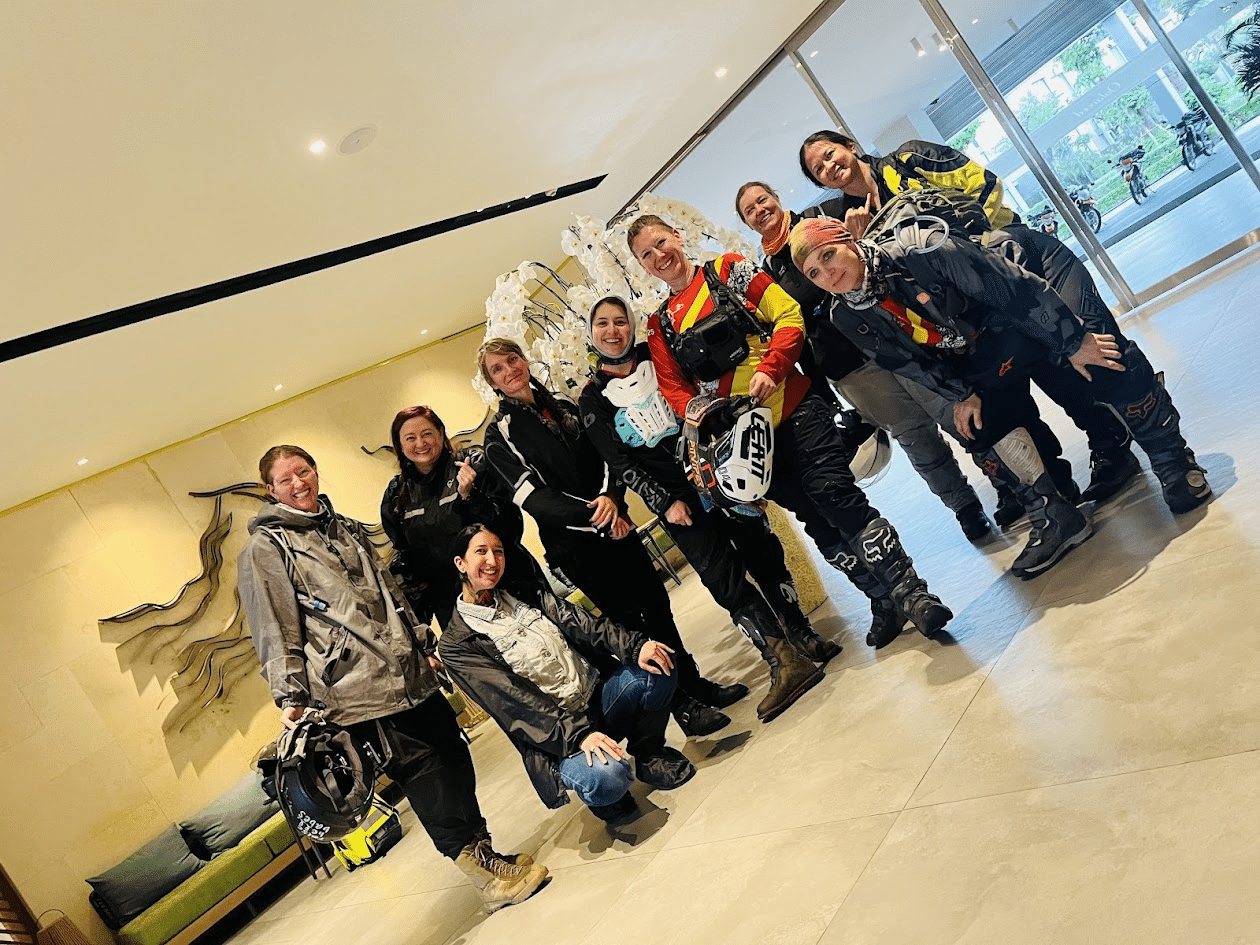 Group of women in motorcycle gear posing indoors near large glass doors and decorative white flowers.