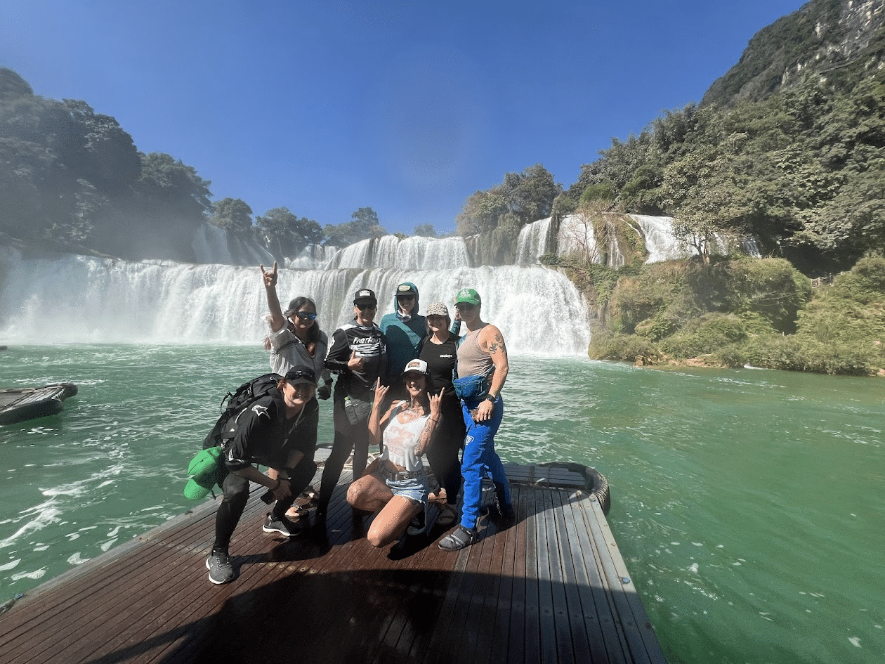 Group of tourists posing on a wooden platform near a large cascading waterfall with lush greenery and clear blue sky.