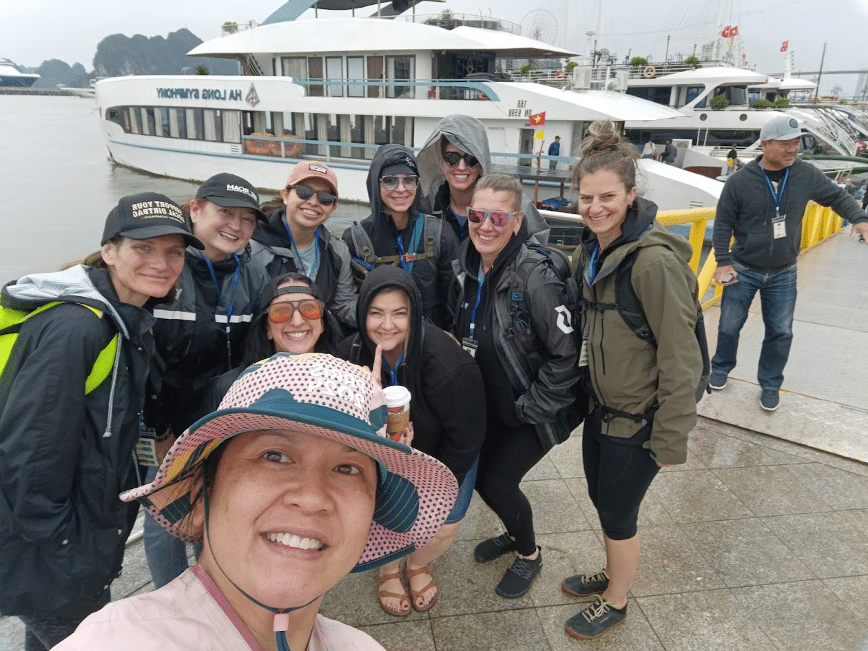 Group of tourists smiling and posing for a selfie on a dock with a white cruise boat in the background.