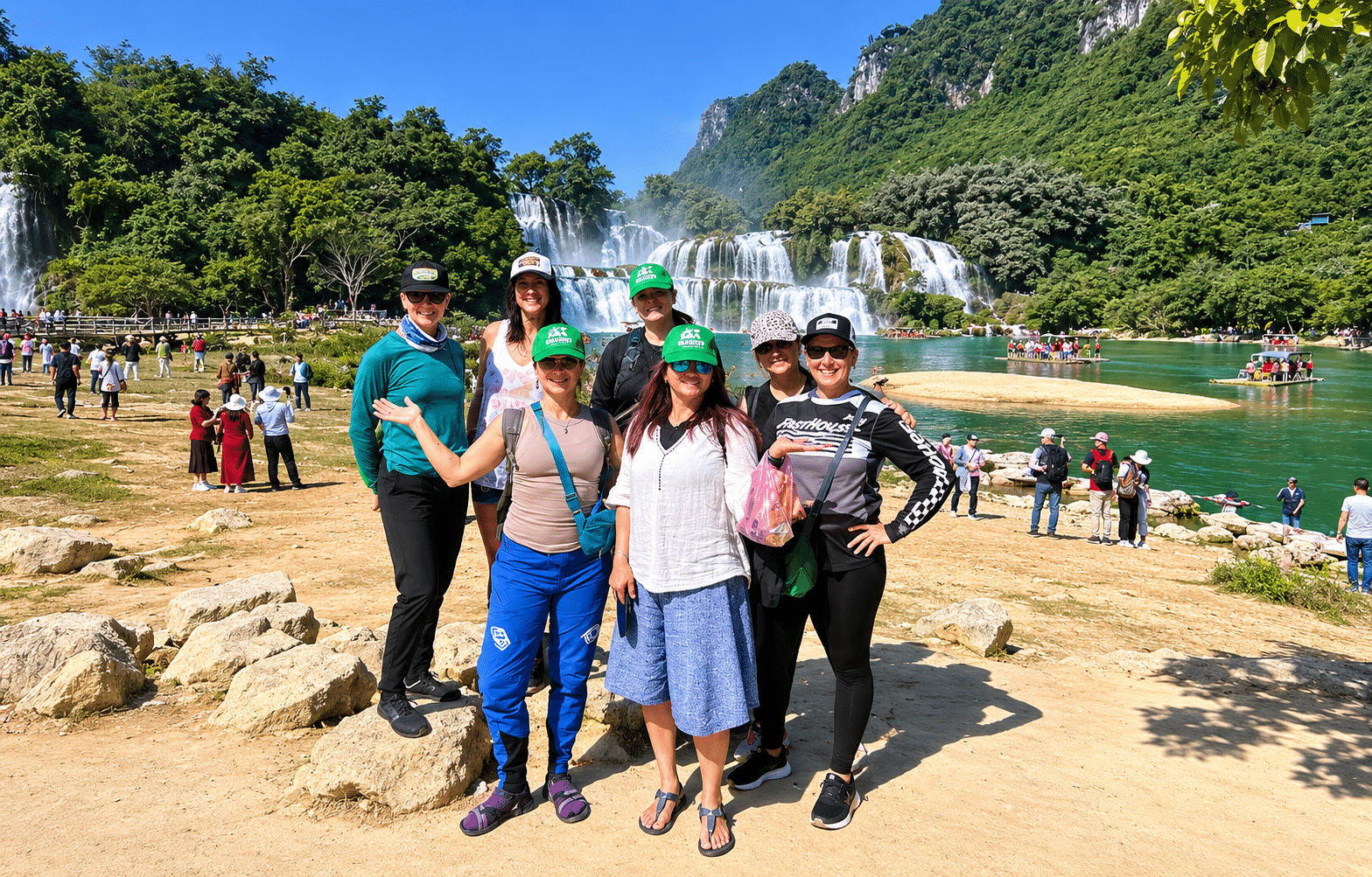 Group of tourists posing near vibrant green Bach Ma Waterfall in Ban Gioc, Vietnam on a sunny day.