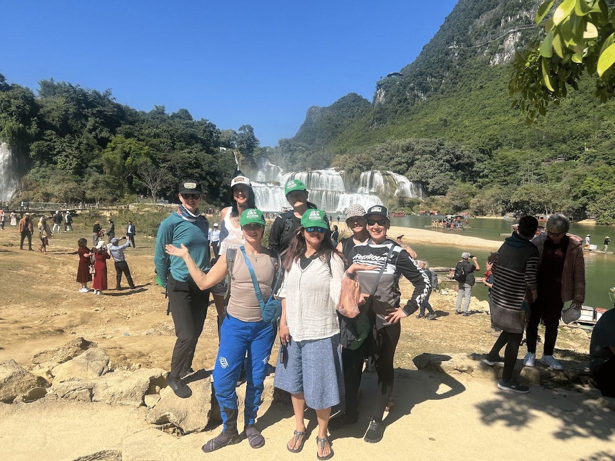 Group of tourists posing in front of a large waterfall and lush green mountains on a sunny day.