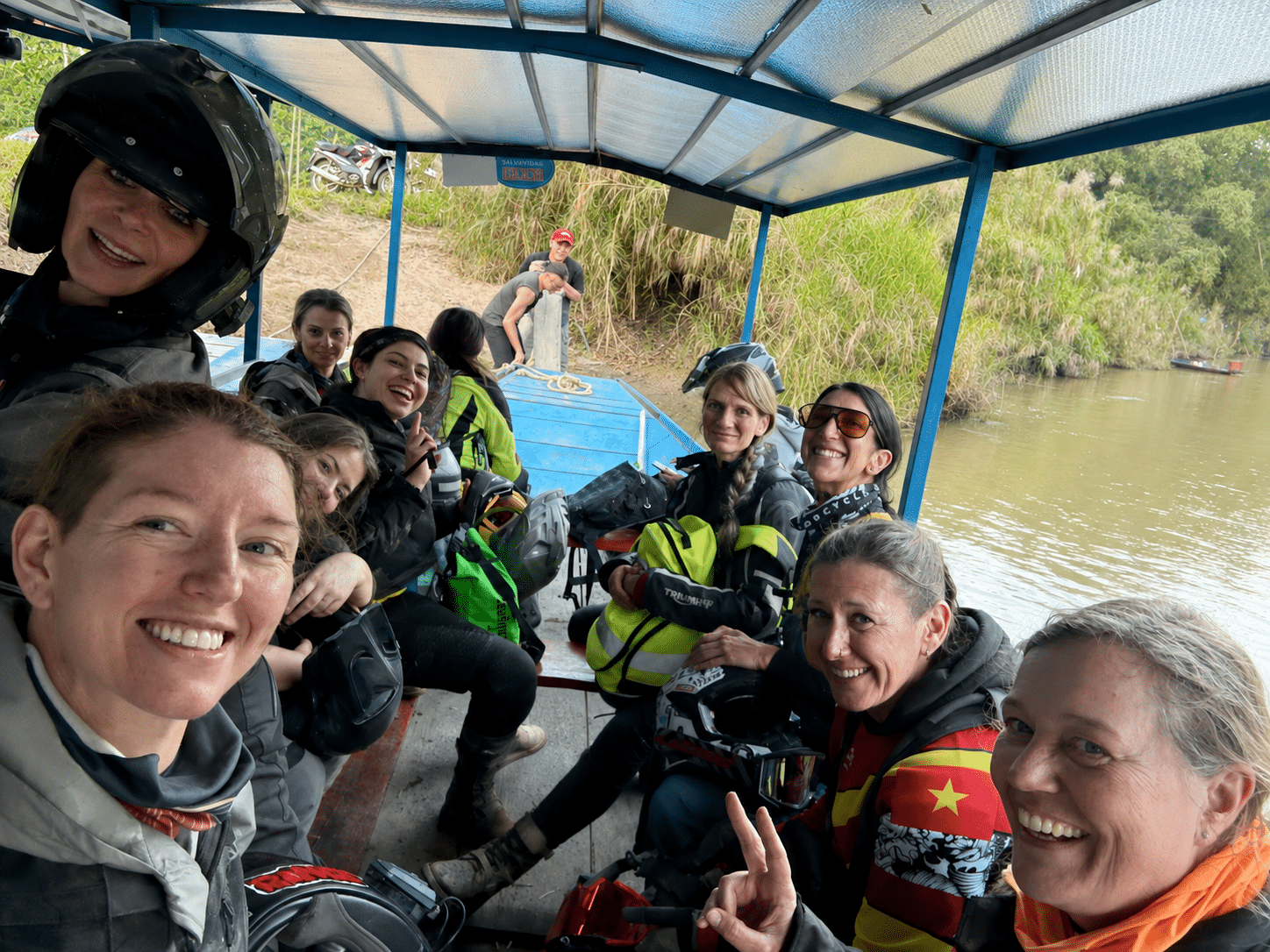 Group of smiling women wearing motorcycle gear on a covered boat ride along a river with green vegetation in the background.