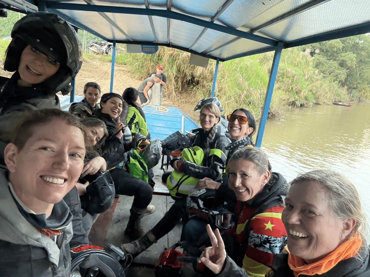 Group of smiling motorcyclists wearing helmets and gear on a covered boat crossing a river with greenery in the background.