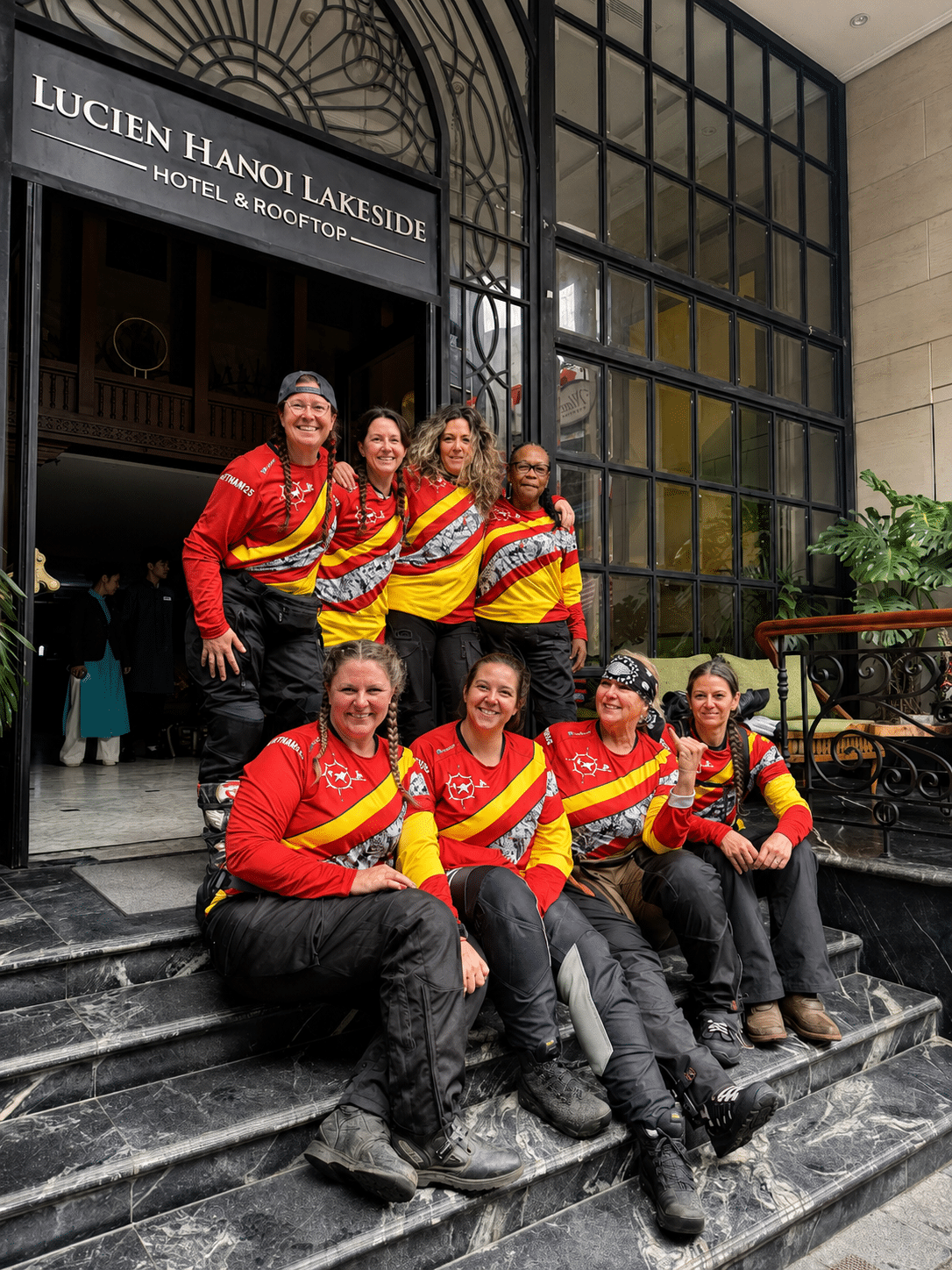 A group of seven women wearing bright red and yellow uniforms poses together on the steps of a building, showcasing teamwork and camaraderie. The setting appears to be a public venue, with greenery in the background.