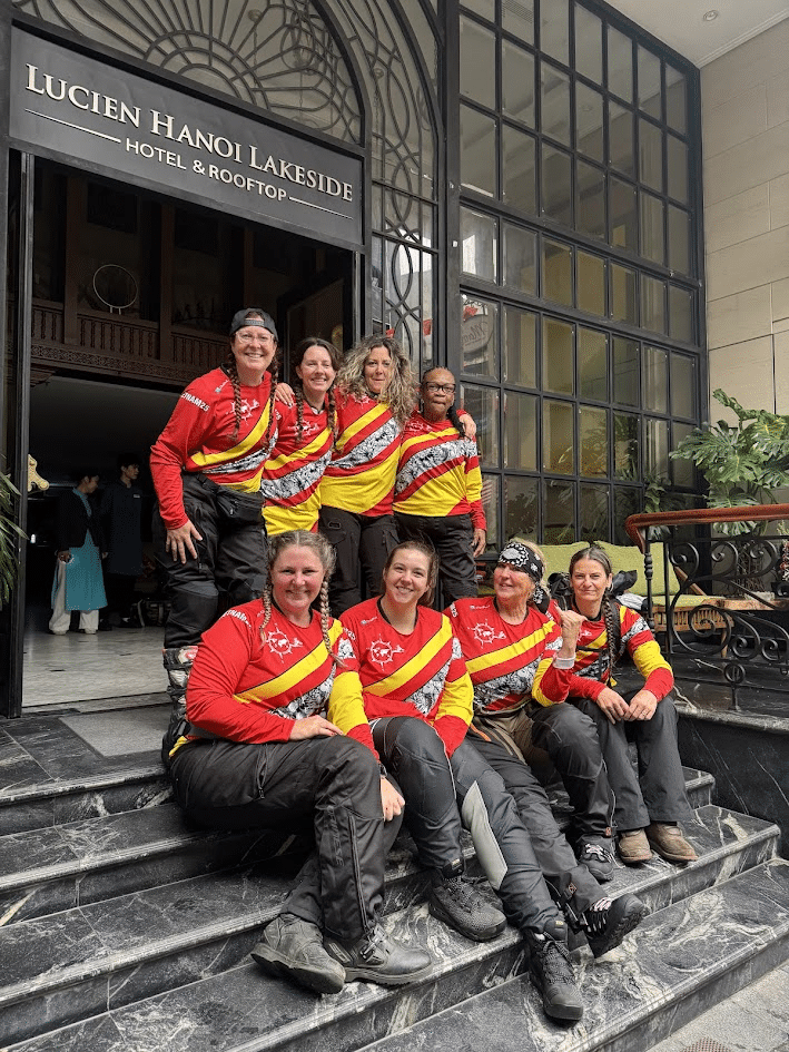A group of seven women wearing bright red and yellow uniforms poses together on the steps of a building, showcasing teamwork and camaraderie. The setting appears to be a public venue, with greenery in the background.