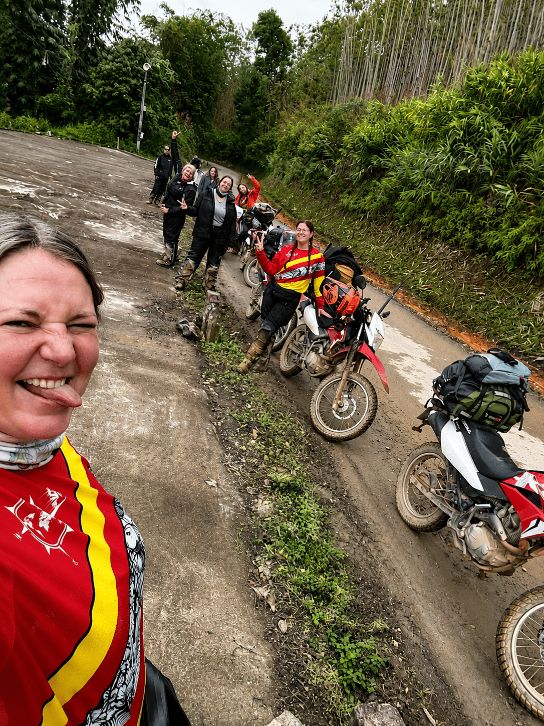 Group of female motorcyclists posing for a selfie on a forest road with dirt bikes and green foliage background.