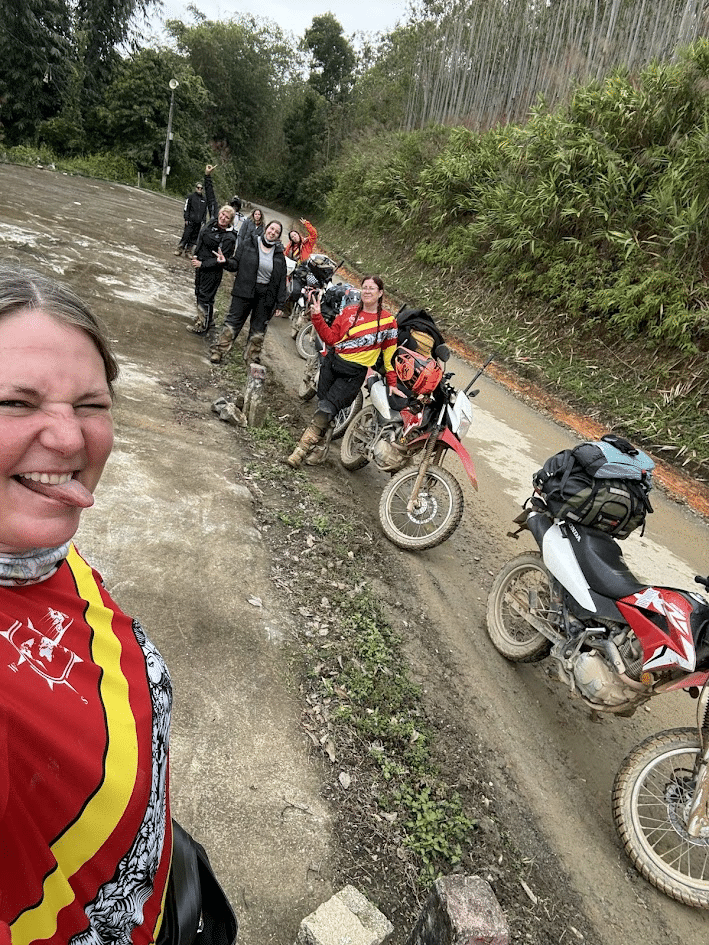 Group of motorcyclists with dirt bikes posing for a selfie on a dirt road surrounded by greenery.