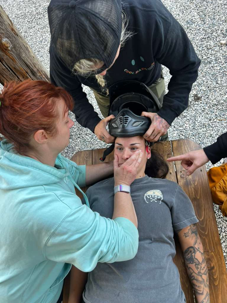 Group of four individuals assisting a person lying on a wooden surface, while carefully placing a helmet on their head. The setting appears to be outdoors, with gravel and natural elements in the background.