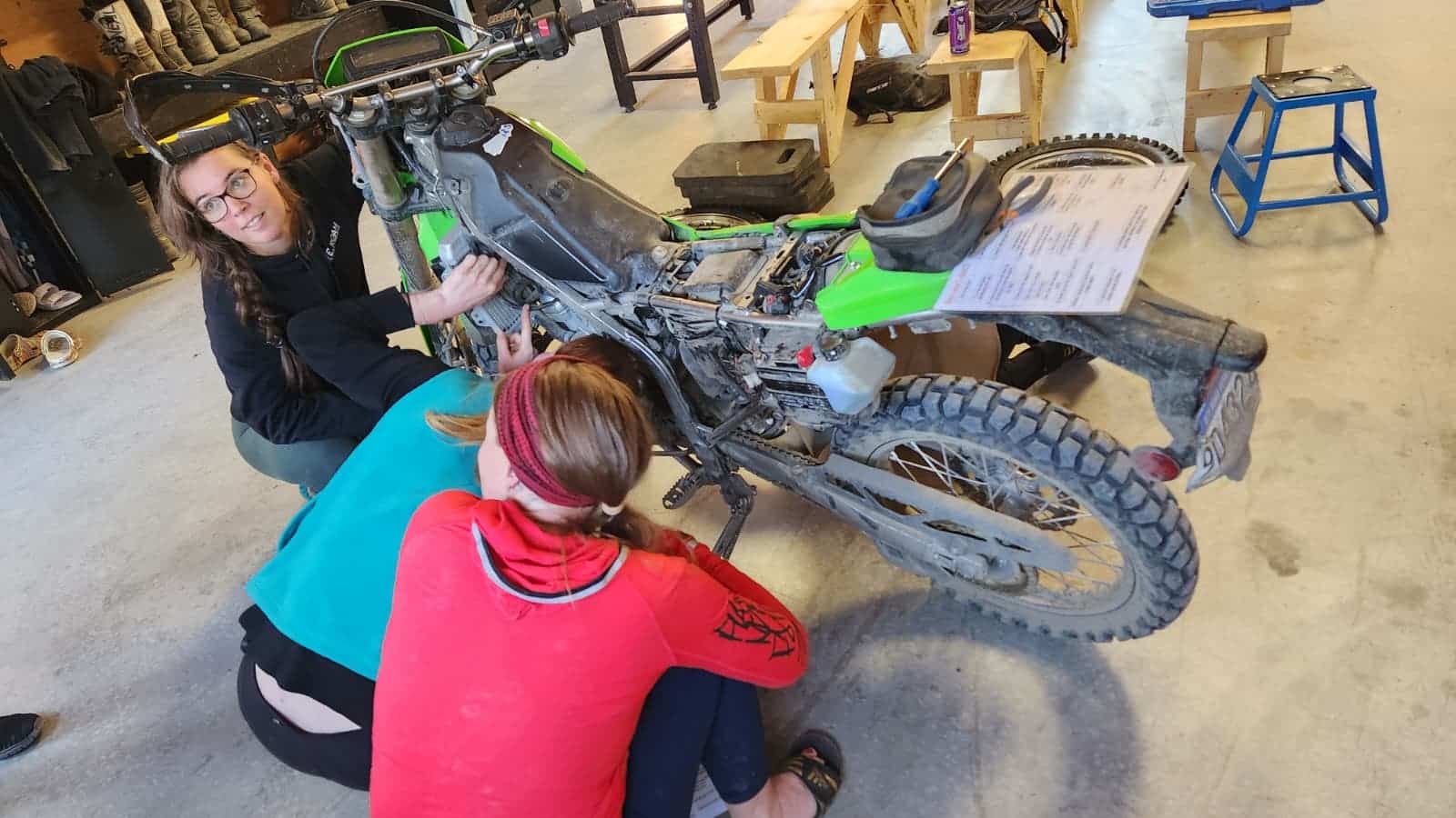 A person in a red and black outfit is working on a dirt bike in a garage setting, focusing on maintenance and repairs. The bike is green and has visible dirt and wear, indicating recent use. Tools and equipment are scattered around, emphasizing a hands-on approach to motorcycle upkeep.