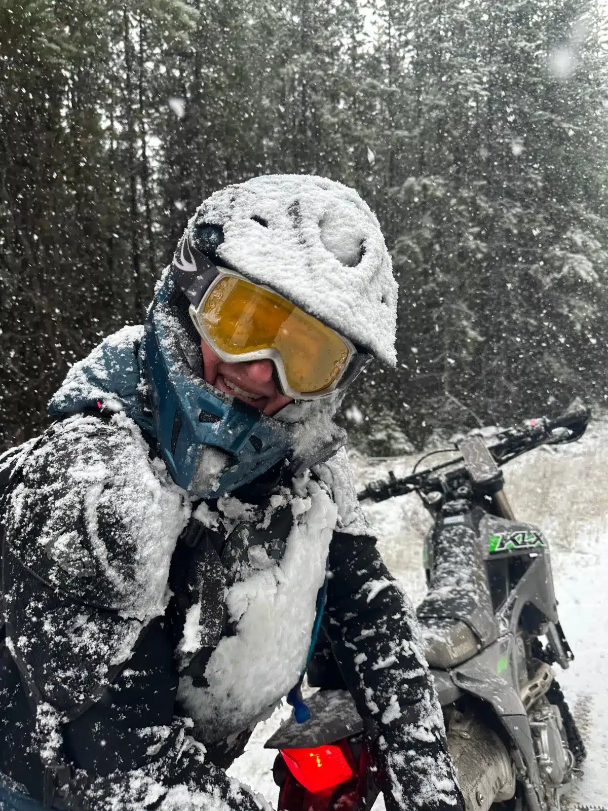 Snowmobile rider in winter gear, covered in snow, posing amidst a snowy forest backdrop.
