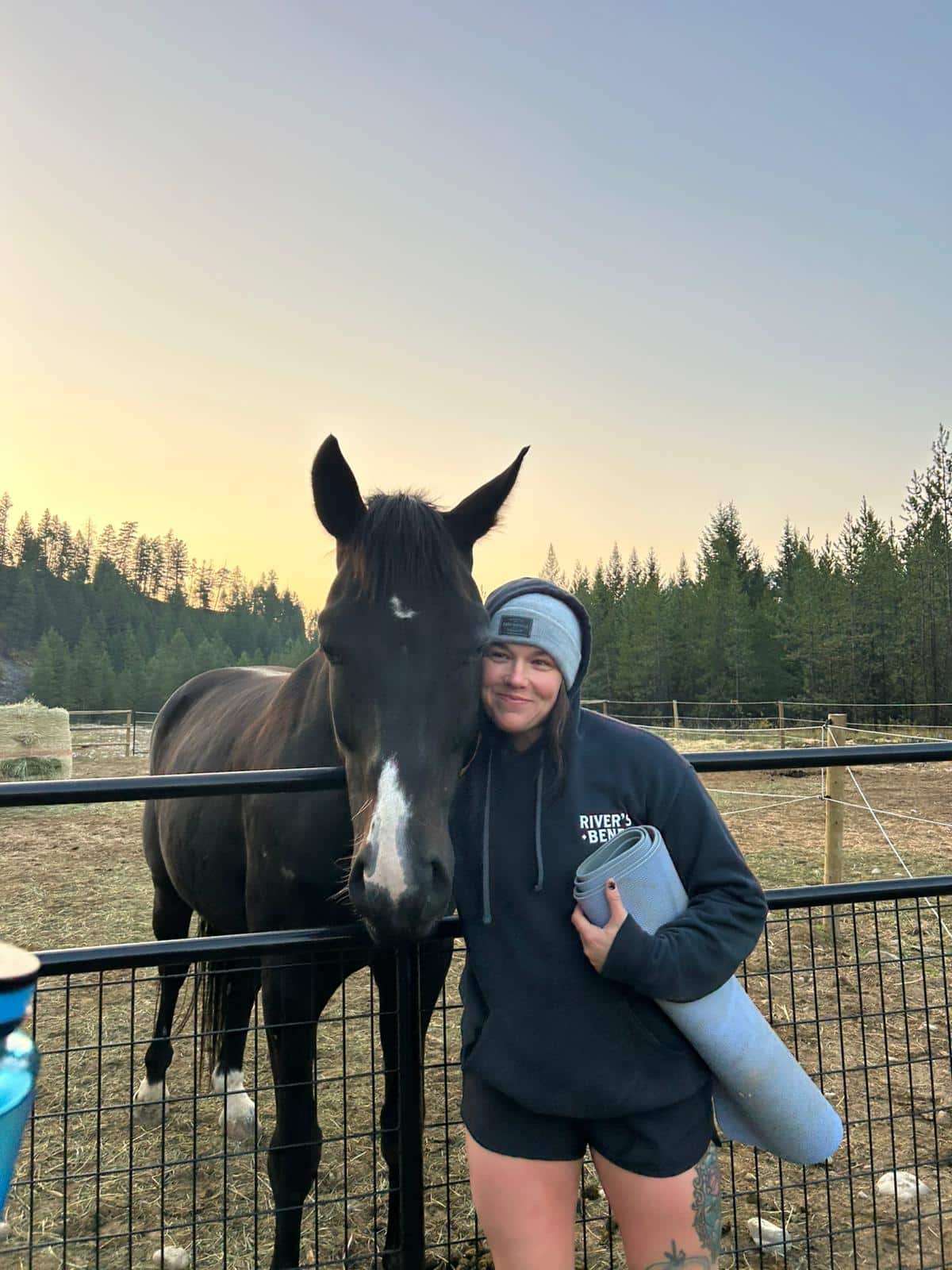 Woman in hoodie and beanie holding a yoga mat, standing next to a black horse by a fence during sunset.