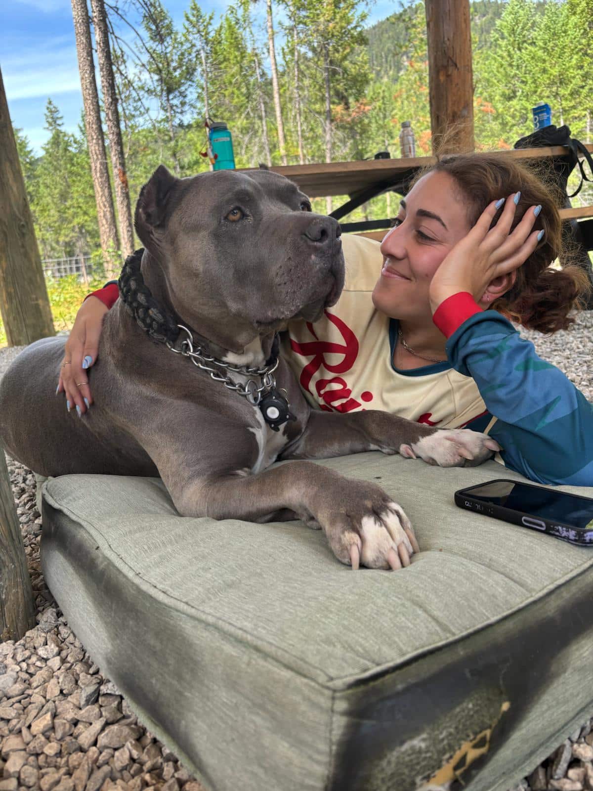 Woman smiling and cuddling with a large gray dog lying on a cushion outdoors in a forested area.
