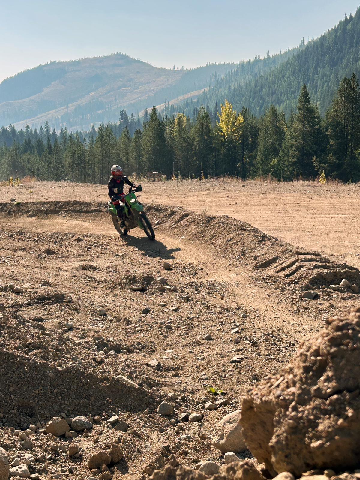 Woman riding a green dirt bike on a dirt trail in a mountainous forest area on a sunny day.