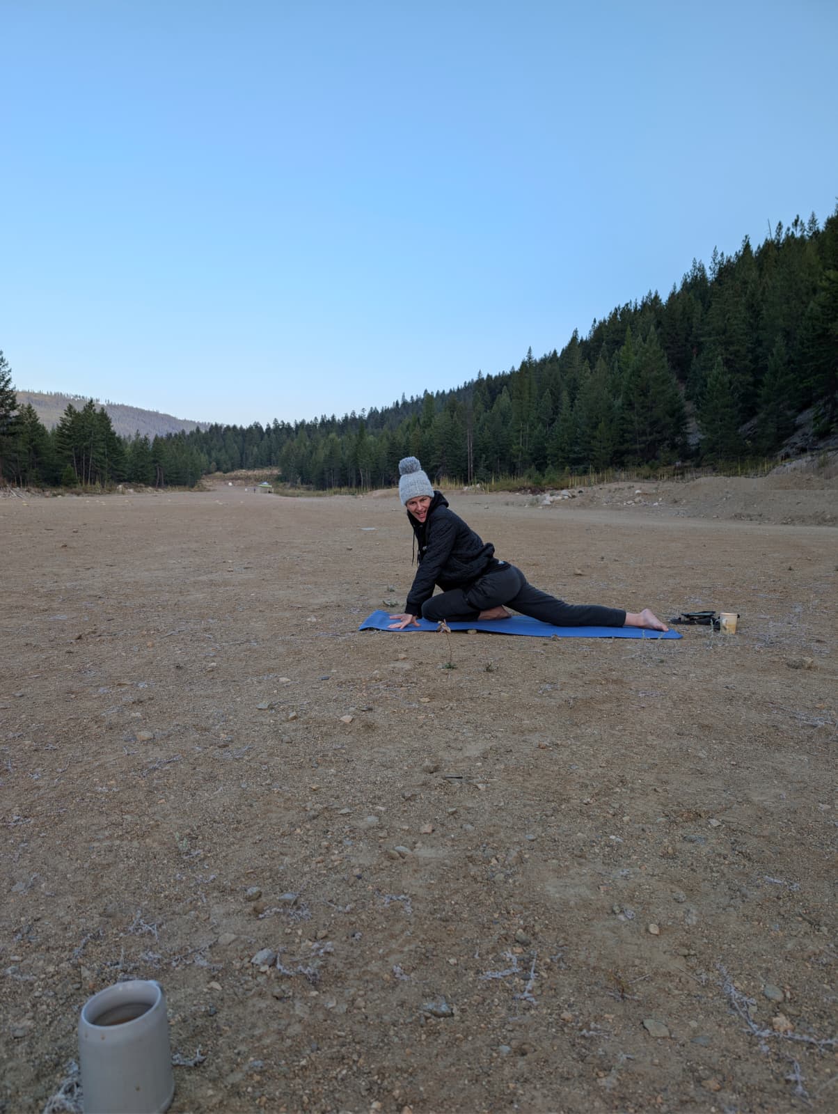 Woman practicing yoga on a blue mat in a wide open dirt area surrounded by pine trees under a clear blue sky.